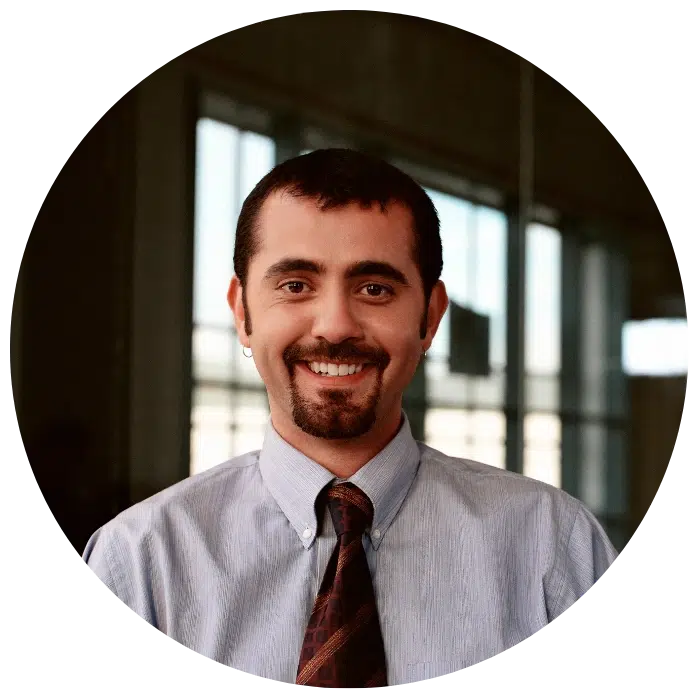 A man with short dark hair, a goatee, and an earring smiles at the camera. He is wearing a light blue dress shirt and patterned tie, standing indoors with windows in the background.